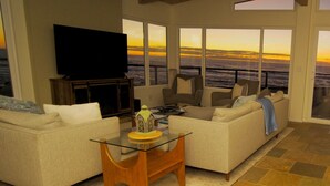 Ein geräumiges Wohnzimmer mit einem großen Flachbildfernseher, bequemen Sitzgelegenheiten und Meerblick bei Sonnenuntergang.