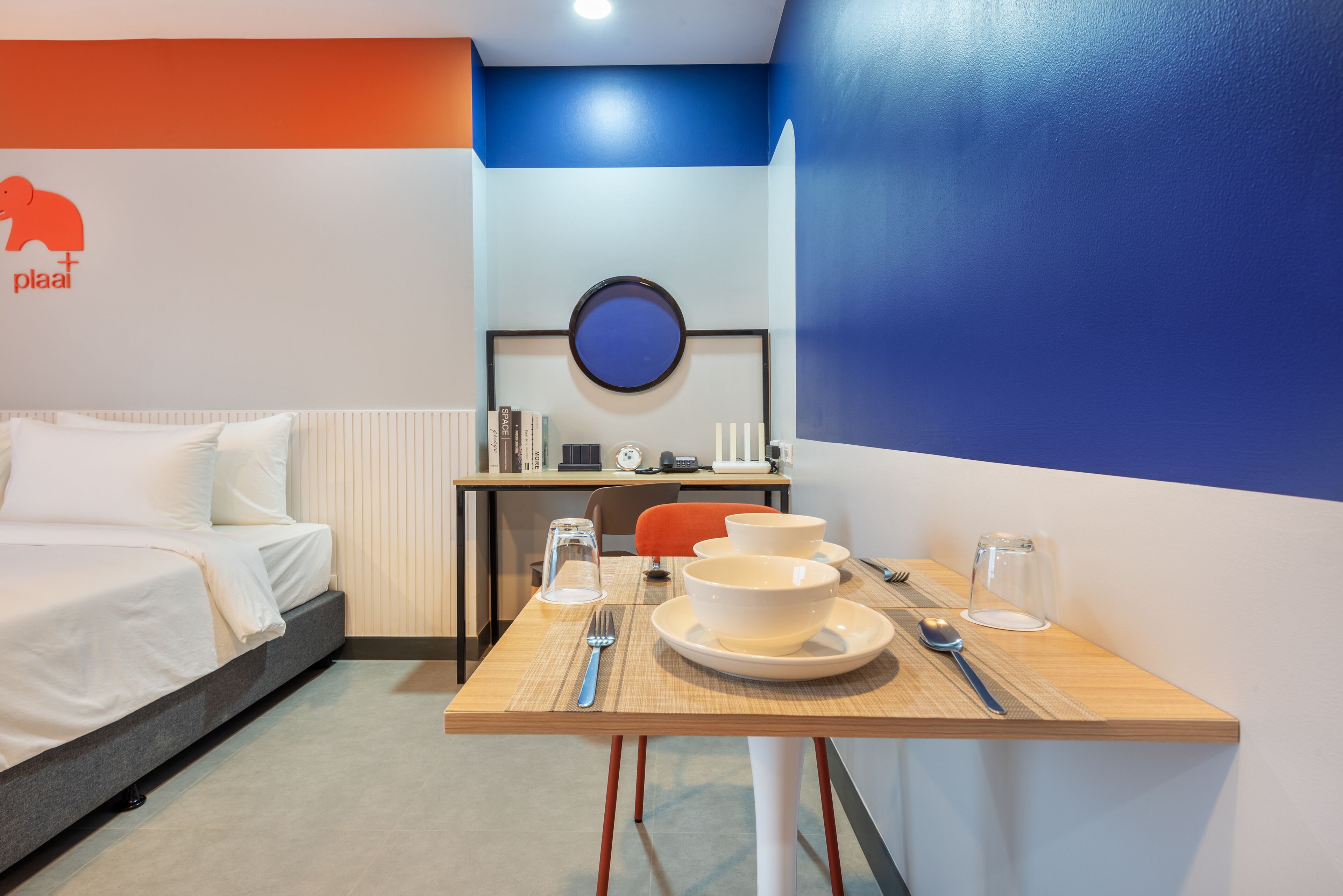 A hotel room with a bed, a desk with a chair, a dining table with a bowl and cutlery, and a wall with a logo.