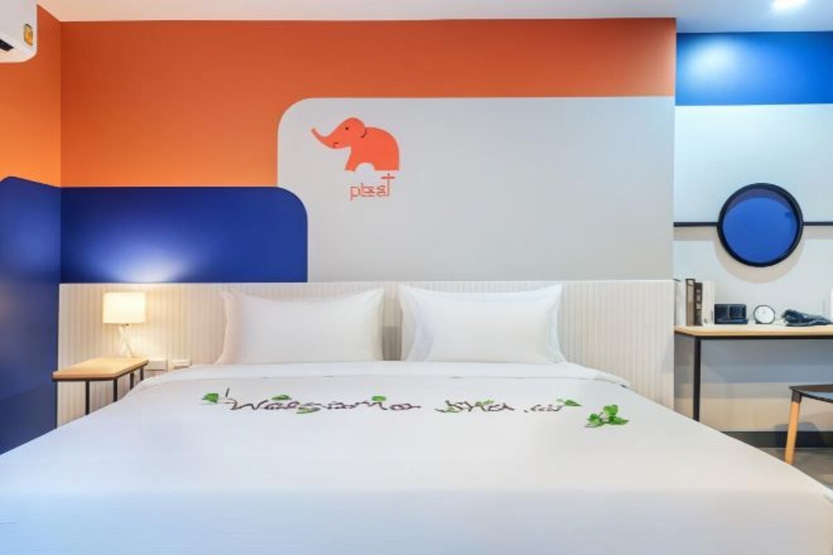 A hotel room with a bed, a nightstand, a lamp, a desk, and a wall with a logo and text.