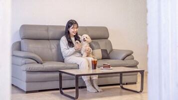 Una persona sentada en un sofá gris, con un perro pequeño, sosteniendo un premio y un vaso de té helado sobre una mesa de centro.