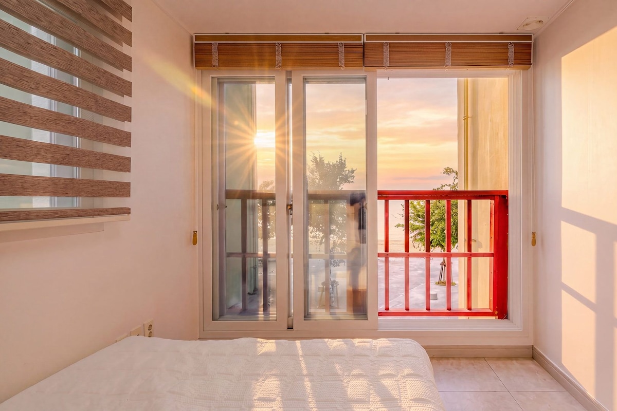 Un dormitorio con cama, una puerta corrediza de vidrio que da a un balcón con barandilla roja y vistas al atardecer.