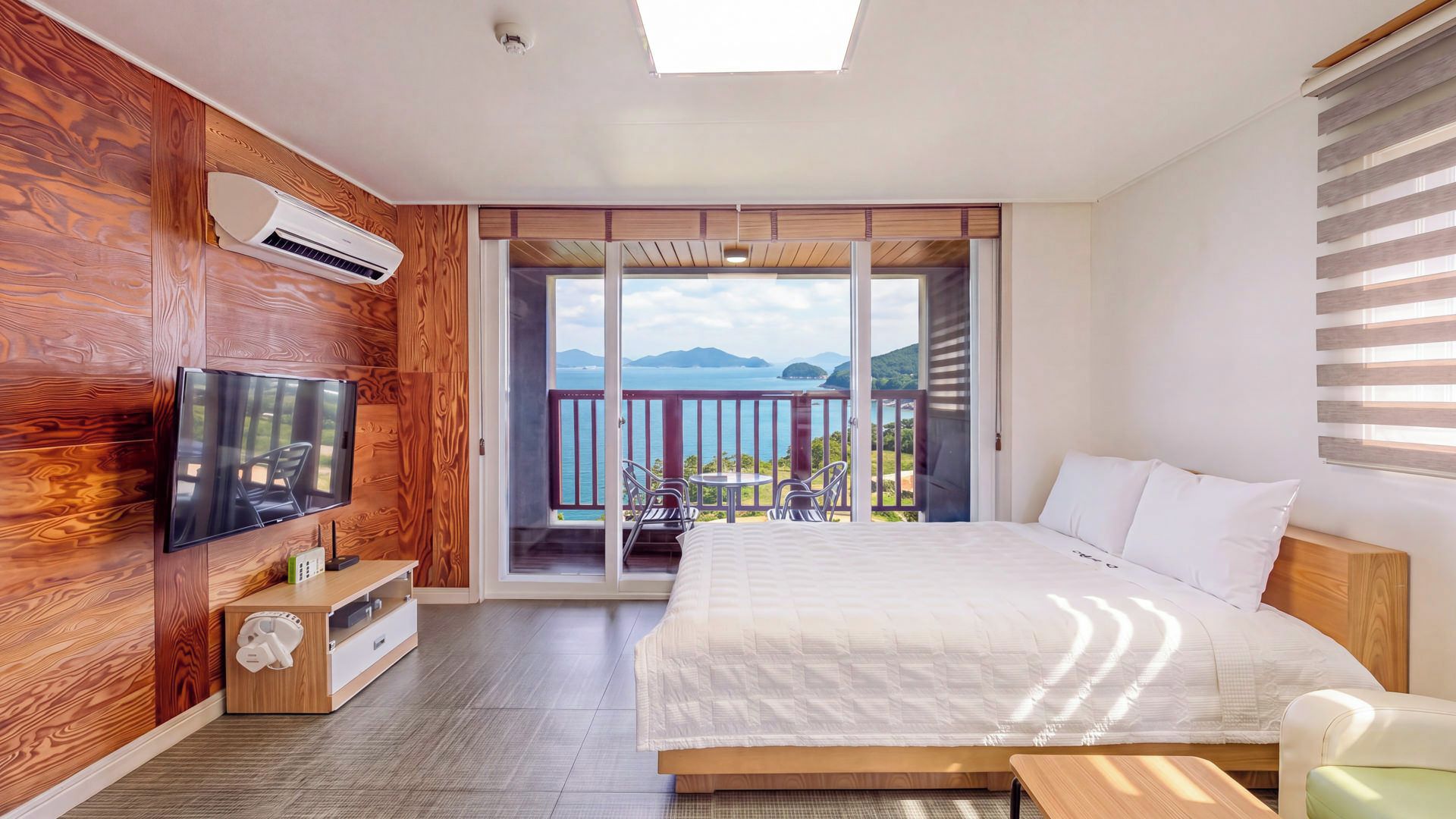 A bedroom with a large bed, a TV mounted on the wall, a wooden floor, and a balcony with a view of the sea.