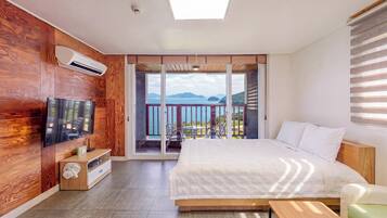 A bedroom with a large bed, a TV mounted on the wall, a wooden floor, and a balcony with a view of the sea.