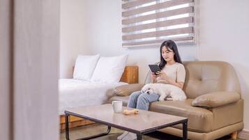 A person sitting on a beige sofa with a white dog, using a tablet. There is a white bed with a wooden headboard in the background, and a small table with a mug and a bone-shaped object on it.