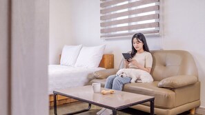 Una persona sentada en un sofá color crema con un perro blanco, usando una tableta. En el fondo, hay una cama blanca con cabecera de madera, y una mesita con una taza y un objeto con forma de hueso.