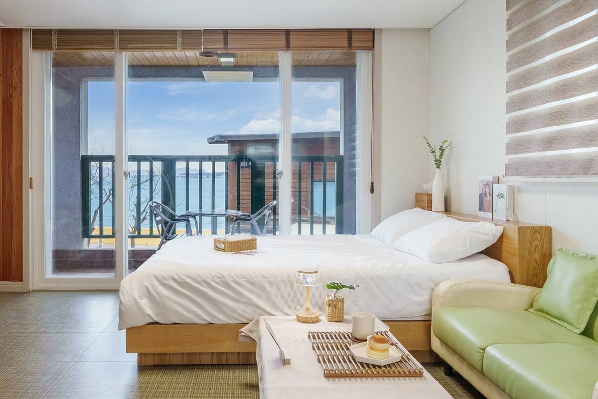 A modern hotel room with a large sliding glass door leading to a balcony, a bed with white linens, a green sofa, and a small table with a tray of food.