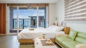 Una habitación de hotel moderna con un amplio ventanal corredera que da a un balcón, una cama con ropa de cama blanca, un sofá verde y una pequeña mesa con una bandeja de comida.