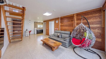 Una sala moderna con una escalera de madera, un sofá gris, un sillón colgante y una cocina al fondo.