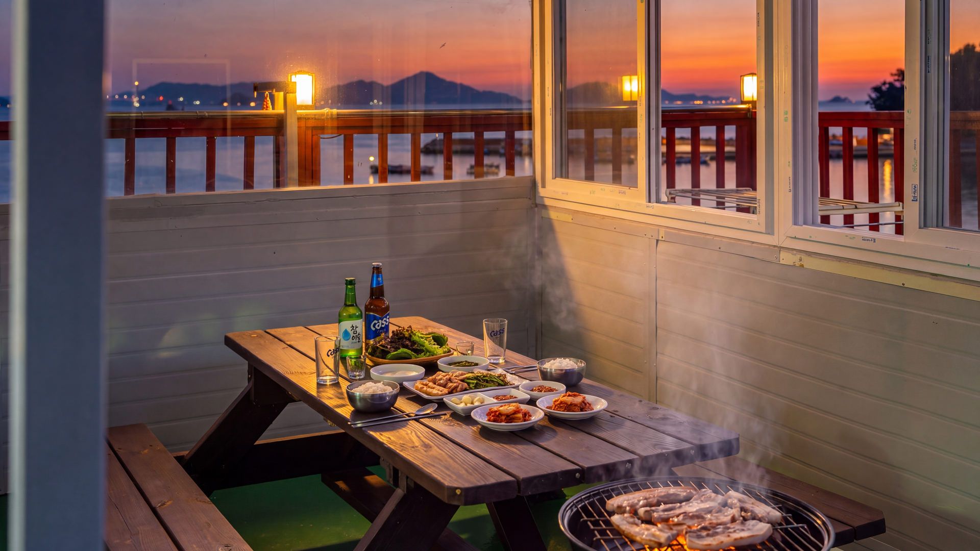 A wooden table set with food and drinks, a grill with meat, and a view of the sunset over water.