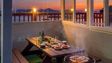 A wooden table set with food and drinks, a grill with meat, and a view of the sunset over water.