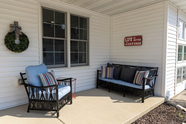 A porch with two black wicker chairs and a blue cushioned bench, a white exterior wall, and a wreath with a bow.