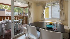 Uma área de refeições ao ar livre coberta, com mesa e cadeiras, uma janela com cortinas e vista para um deck de madeira e uma casa.