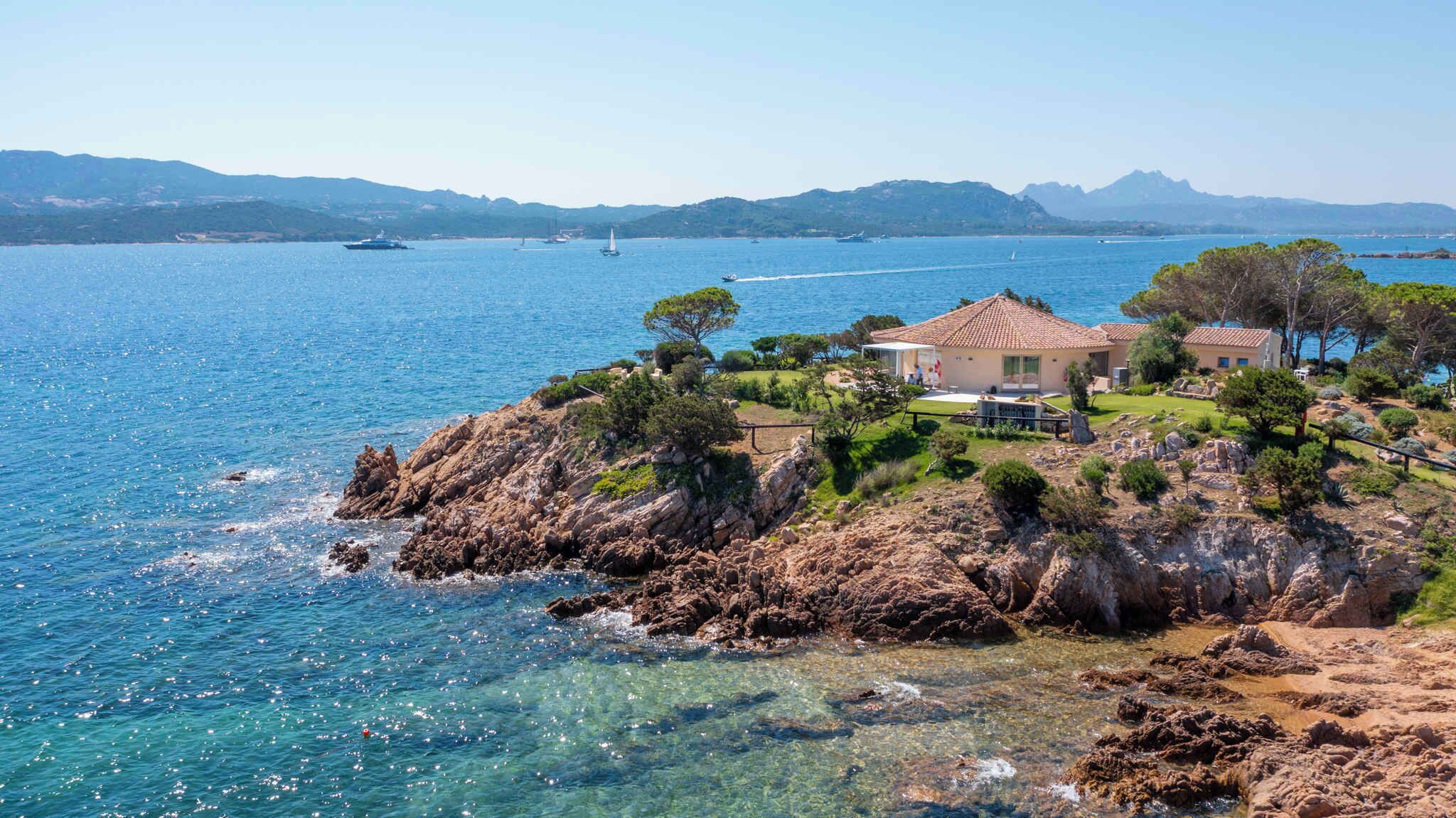 Uma propriedade costeira com uma casa em um afloramento rochoso, mar azul límpido e barcos ao longe.