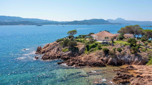 A coastal property with a house on a rocky outcrop, clear blue sea, and boats in the distance.