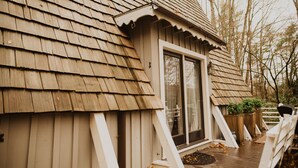 A wooden house with a shingled roof, a sliding glass door, and a deck with a white railing.