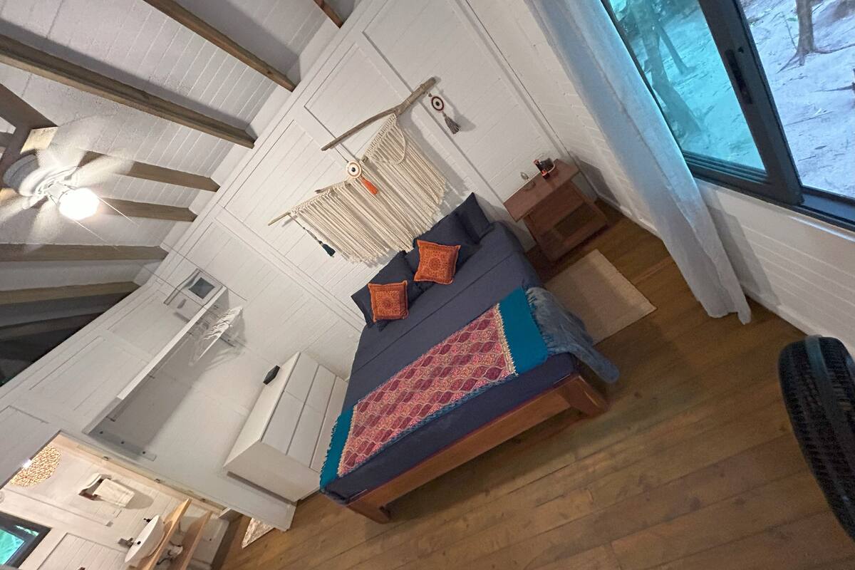 Ein Schlafzimmer mit einem Bett, einem Deckenventilator, einer Wanddekoration aus Makramee und einem Fenster mit Blick auf Bäume.