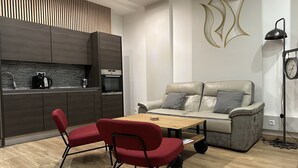 Une cuisine moderne avec des meubles en bois foncé, un four encastré et une machine à café. On y trouve une chaise rouge et une table en bois avec des livres. Un canapé gris est placé contre le mur, et une lampe sur pied est visible.