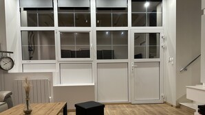 Une pièce moderne avec un plancher en bois, un pouf noir, une table en bois ornée d’un vase et une grande fenêtre à carreaux.