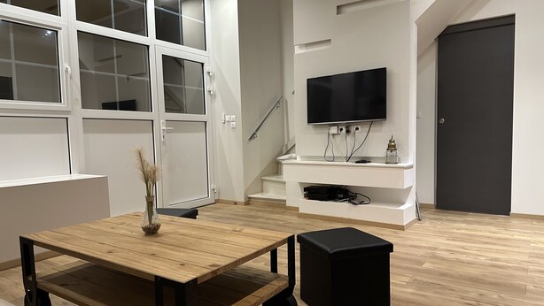 Un salon moderne avec une table en bois, un pouf noir, un téléviseur posé sur un meuble blanc et un escalier menant à un niveau supérieur.