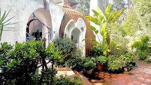 Une cour avec un sol carrelé, des plantes en pot et un bâtiment doté d’un toit et d’arcades couverts de tuiles.