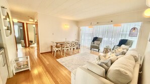 A spacious living area with a wooden floor, a beige sofa, a dining table with chairs, and a large window with sheer curtains. - Pipi Palace Ruakaka – Ideal for Work & Family Stays (Ruakaka)