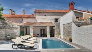 Un espace aménagé au bord de la piscine, avec des chaises longues, une piscine, des murs en pierre et un toit recouvert de tuiles rouges.