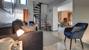 Un salon moderne avec un canapé foncé, un fauteuil bleu et un escalier en colimaçon menant à un niveau supérieur.