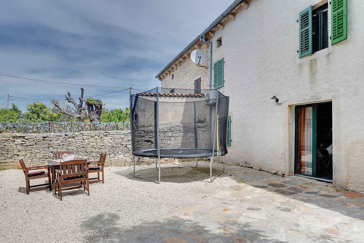 Une terrasse avec un trampoline, un mur en pierre et un bâtiment blanc aux volets verts.
