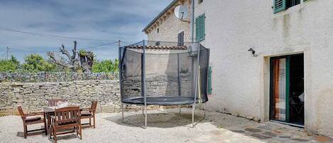 Une terrasse avec un trampoline, un mur en pierre et un bâtiment blanc aux volets verts.