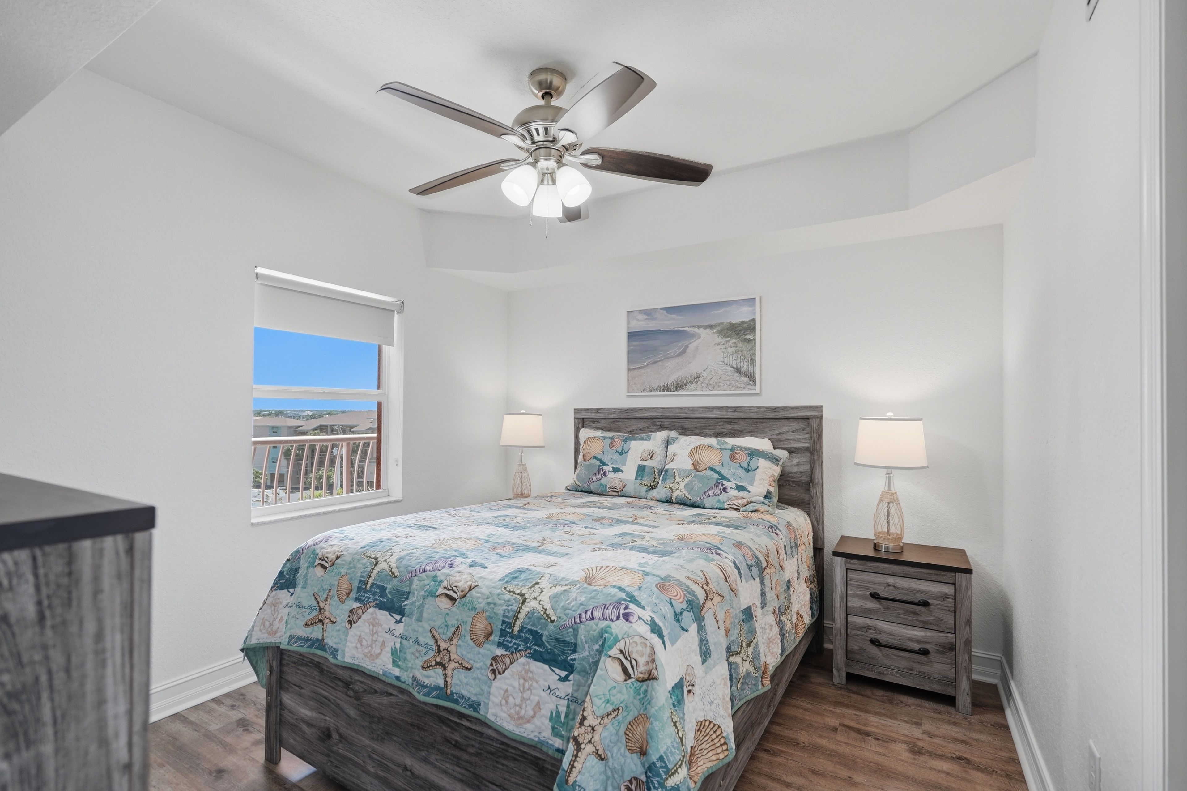A bedroom with a bed, a nightstand, a ceiling fan, a window with a view, and a painting on the wall.