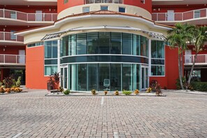 A modern building with a curved glass facade, multiple balconies, and a paved entrance area with potted plants.