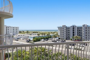 A balcony with a view of a parking lot, a beach, and a multi-story building.