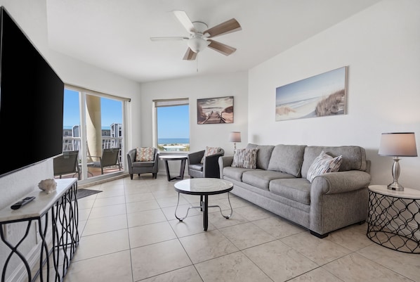 A spacious living room with a grey sofa, a coffee table, and a TV. There are two windows with a view of the sea.