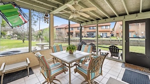 Ein überdachter Patio-Bereich mit Tisch und Stühlen, einem Deckenventilator und großen Fenstern mit Blick auf die umliegende Nachbarschaft.
