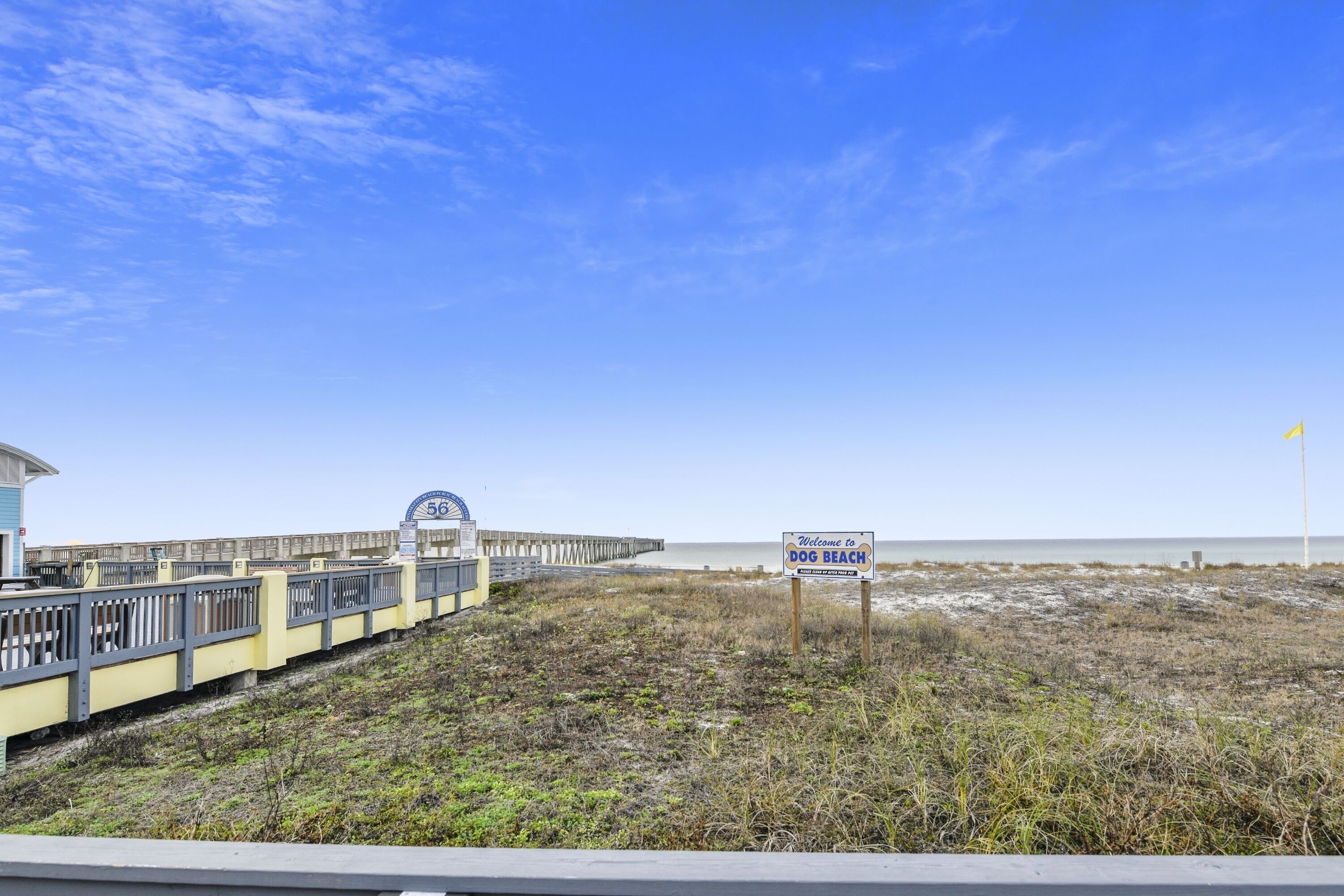 A beach with a wooden boardwalk, a sign reading "Welcome to Dog Beach," and a pier in the background.