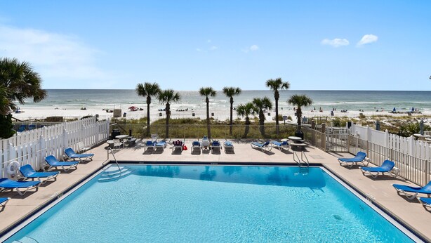 Ein Pool mit blauen Liegestühlen, einem weißen Zaun und Blick auf Strand und Meer.