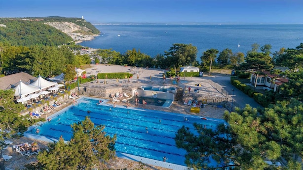 Une piscine dotée de plusieurs couloirs, entourée de chaises longues et de parasols, et offrant une vue sur la mer et les collines en arrière-plan.