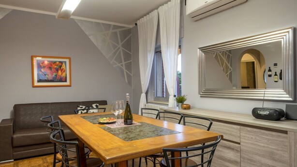 A dining area with a wooden table, chairs, a sofa, a painting, a window with curtains, and a mirror with wine bottles.