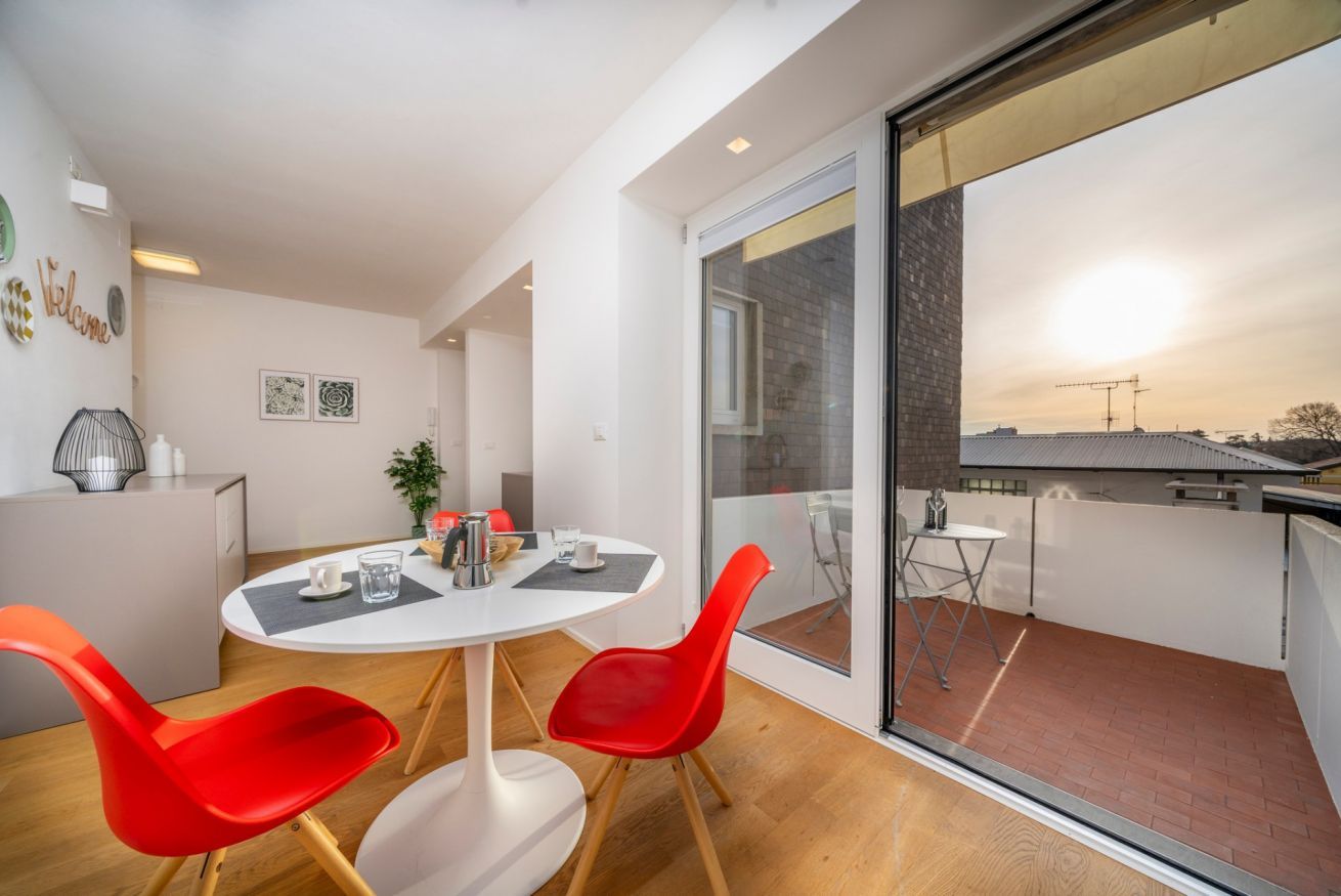 A modern interior with a round dining table, red chairs, a small kitchen area, and a balcony with a view of the cityscape.