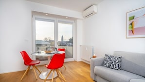 A modern living room with a grey sofa, red chairs, a round table, and a large window with a view of buildings. - GuestHost - Cozy & Bright Apartment - w/ Balcony (udine)