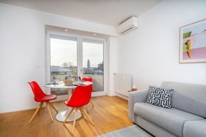 A modern living room with a grey sofa, red chairs, a round table, and a large window with a view of buildings.
