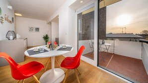 A modern interior with a round dining table, red chairs, a small kitchen area, and a balcony with a view of the cityscape. - GuestHost - Cozy & Bright Apartment - w/ Balcony (udine)