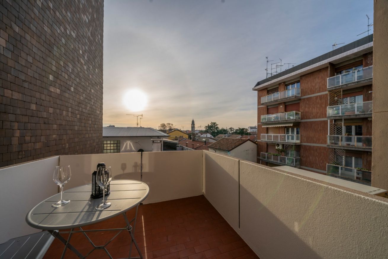 Un balcon avec une table et deux verres à vin, donnant sur un paysage urbain avec des bâtiments et un ciel dégagé.