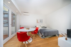 Une chambre moderne avec un lit, une table ronde entourée de chaises rouges, un téléviseur et une vue sur l’extérieur, visible à travers une porte vitrée.