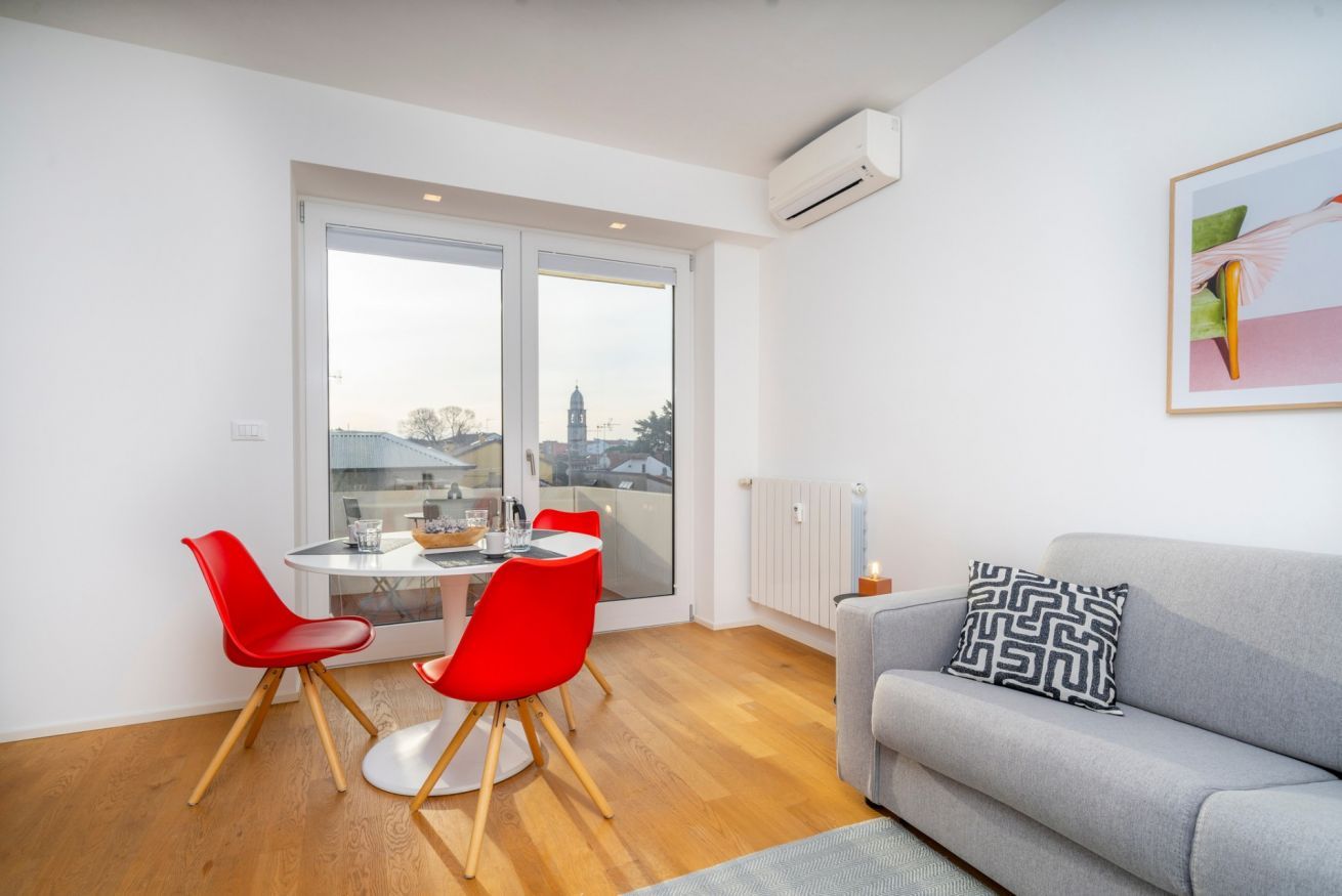 Ein modernes Wohnzimmer mit einem grauen Sofa, roten Sesseln, einem runden Tisch und einem großen Fenster mit Blick auf Gebäude.