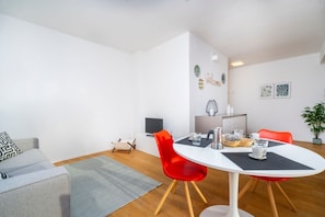 Un salon moderne avec un canapé gris, une table de salle à manger ronde entourée de chaises rouges, une petite télévision et une table basse.