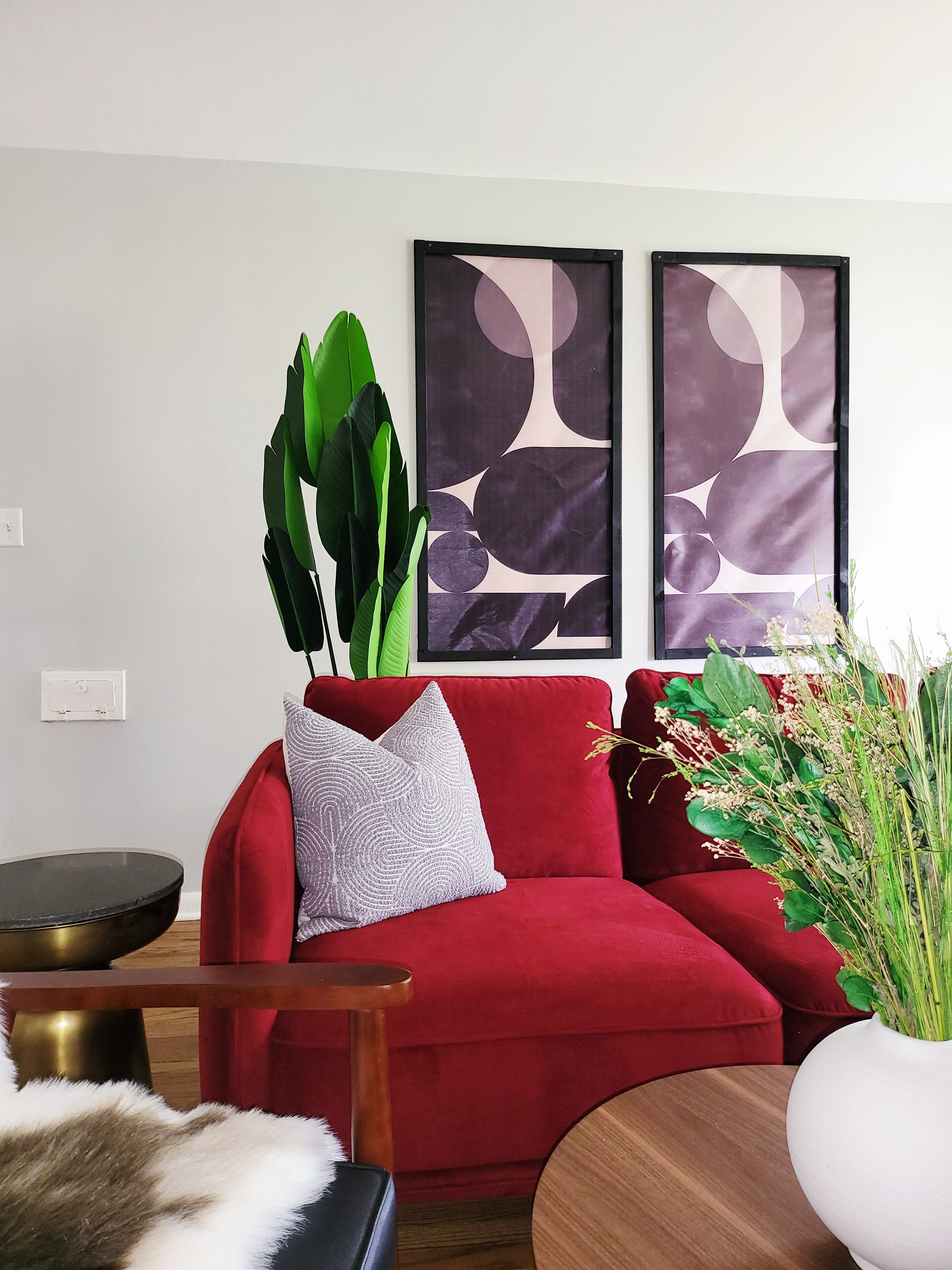Un canapé rouge agrémenté d’un coussin texturé, une grande plante verte et un vase rempli de fleurs. Deux tableaux abstraits sont accrochés au mur.