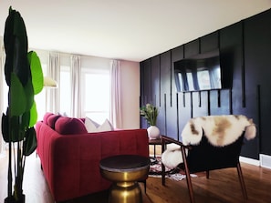 Ein modernes Wohnzimmer mit einem roten Sofa, einem runden goldenen Beistelltisch, einem schwarzen Sessel mit Felldecke, einer großen Pflanze und einem Flachbildfernseher an einer dunklen Wand.