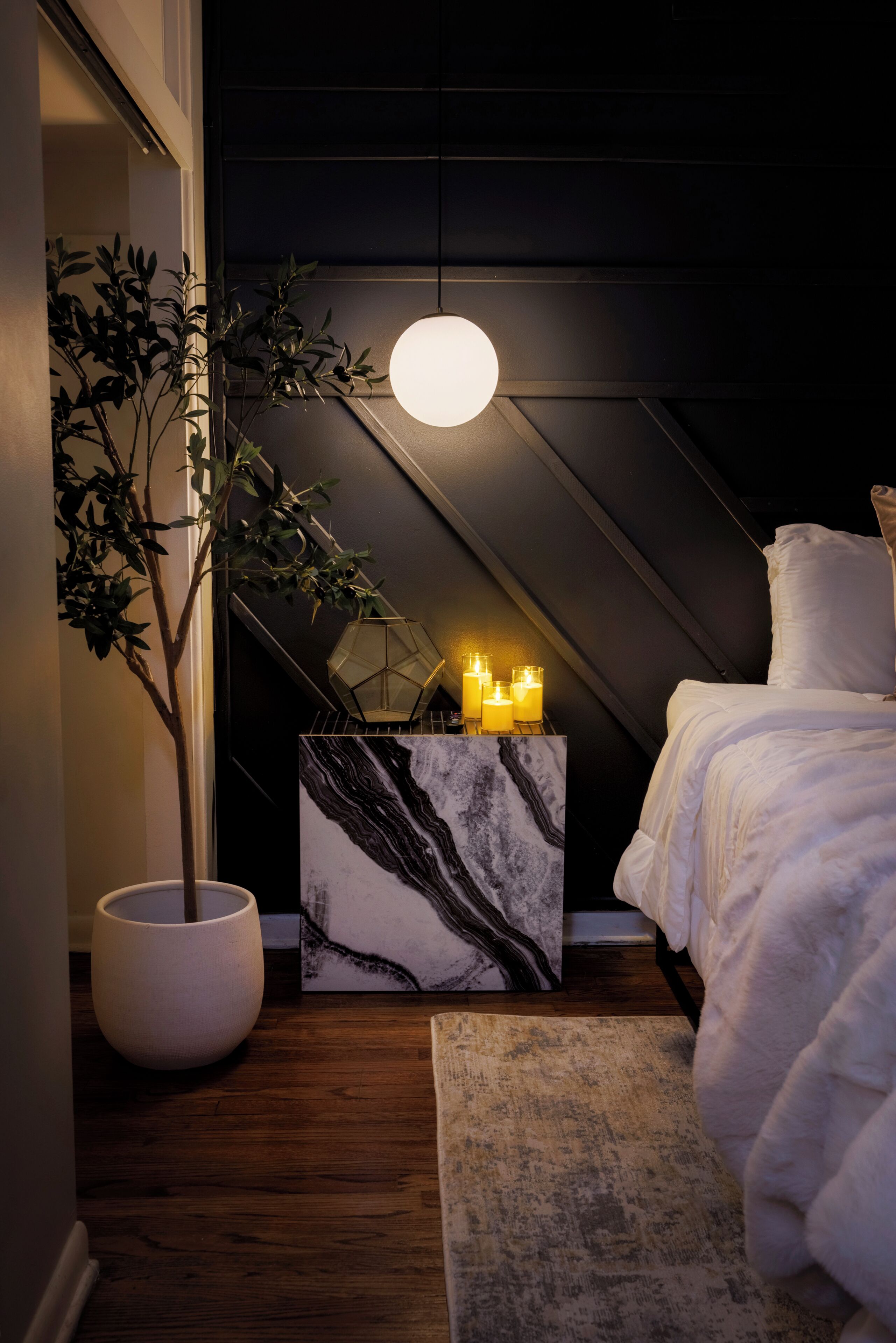 Een slaapkamer met een bed, een potplant, een marmeren bijzettafeltje met kaarsen, en een hanglamp.