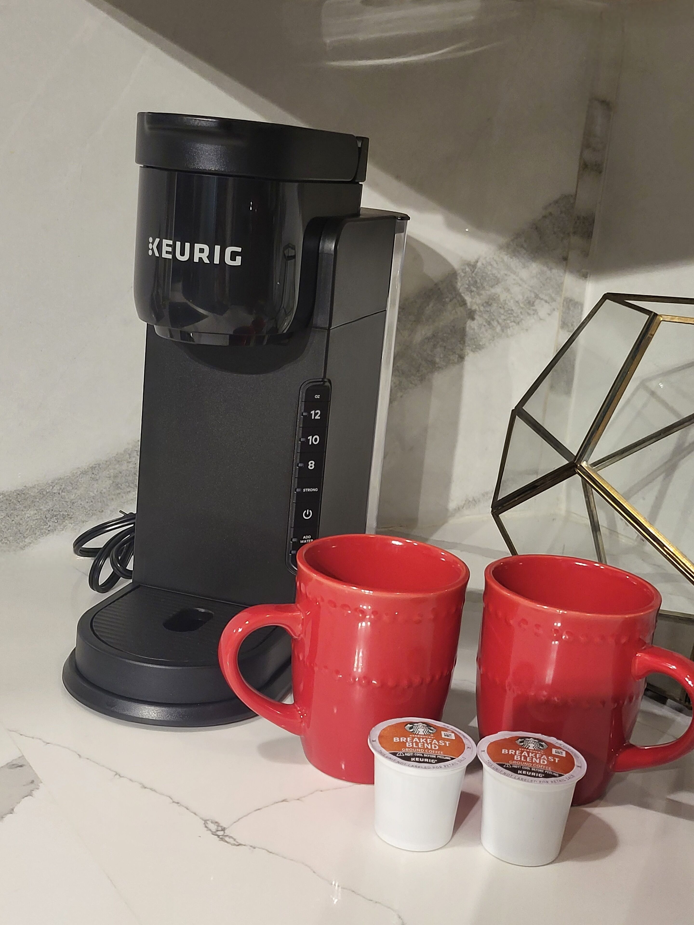 A Keurig coffee maker on a white countertop with two red mugs and two small containers of Keurig coffee pods.
